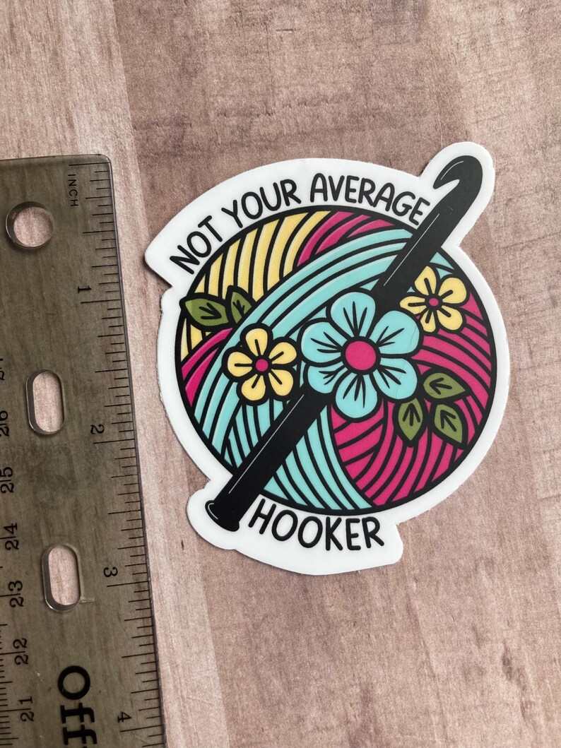 Puede incluir: Una pegatina blanca con una bola de hilo de colores y un gancho de ganchillo negro. El texto en la pegatina dice "Not Your Average Hooker".