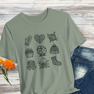 May include: A sage green t-shirt with a black and white graphic design of knitting-themed icons. The icons include a ball of yarn, knitting needles, a mitten, a sheep, a heart, a hat, a basket of yarn, and a person knitting.