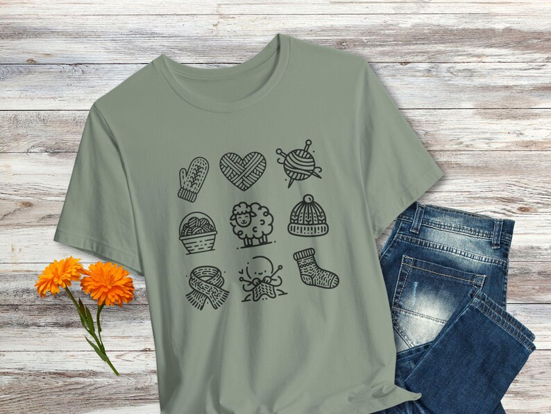 May include: A sage green t-shirt with a black and white graphic design of knitting-themed icons. The icons include a ball of yarn, knitting needles, a mitten, a sheep, a heart, a hat, a basket of yarn, and a person knitting.