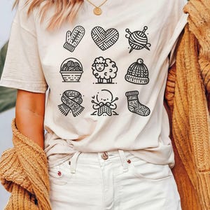 May include: A white t-shirt with a black and white graphic design of knitting-related icons. The icons include a heart, yarn ball, needles, a mitten, a basket of yarn, a sheep, a hat, a sock, and a hand knitting.