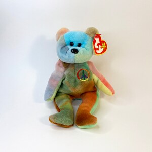 May include: A colorful plush bear with a peace sign on its chest. The bear has a patchwork design with various pastel colors, including blue, pink, green, and brown. It has a red tag attached to its ear.