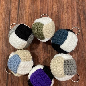 May include: Six crocheted keychains in different colors, each with a different color combination. The keychains are in the shape of a ball and have a silver key ring attached.
