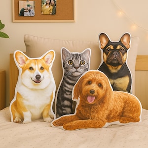 May include: A collection of plush animal pillows in the shapes of a Corgi, a tabby cat, a Goldendoodle, and a black and brown French Bulldog. The pillows are on a bed with a neutral-colored comforter.
