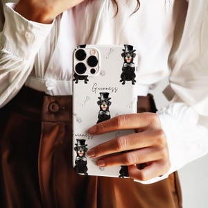 May include: White phone case featuring a repeating pattern of black dogs wearing top hats and bow ties, with bone illustrations. The word "Guinness" is written in cursive. The phone case has a camera cutout.