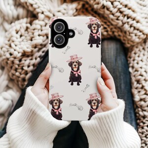 May include: A white phone case featuring a repeating pattern of a black Labrador Retriever wearing a pink hat and bow tie. The background includes sketched bones and rope toys. The phone case is held by a person wearing a white sweater.