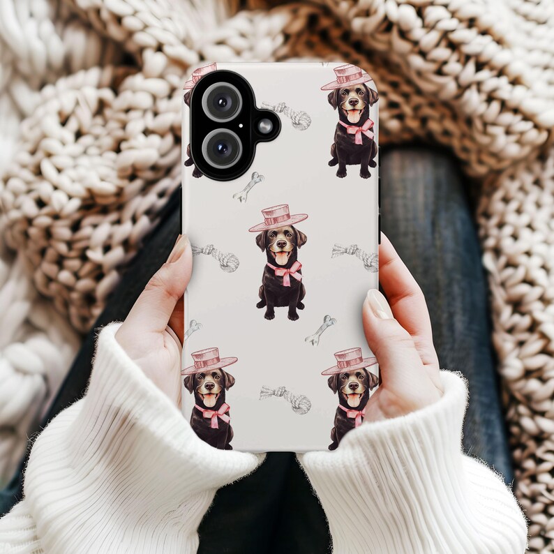 May include: A white phone case featuring a repeating pattern of a black Labrador Retriever wearing a pink hat and bow tie. The background includes sketched bones and rope toys. The phone case is held by a person wearing a white sweater.