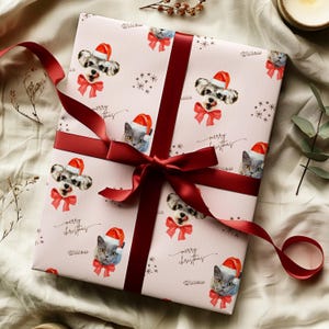 May include: A wrapped gift with light pink wrapping paper featuring a repeating pattern of dogs and cats wearing Santa hats and red bows. The wrapping paper has the words "merry christmas" and "Willow" printed on it. A dark red ribbon is tied around the gift.