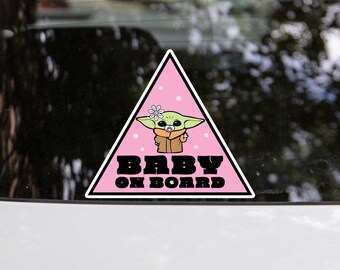 May include: Pink triangular car sticker featuring a cartoon baby Yoda character holding a dummy. The sticker reads "Baby On Board" in black text.