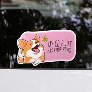 May include: A sticker showing a cartoon corgi with orange and white fur, wearing a flower. The sticker has a pink background with the text "MY CO-PILOT HAS FOUR PAWS". The sticker is attached to a car window.