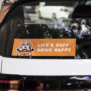 Könnte beinhalten: Orangefarbener und weißer Autoaufkleber mit einem Cartoon-Hund, der ein Auto fährt. Der Aufkleber lautet "Life's Ruff Drive Happy" mit einer kleinen Blumengrafik.