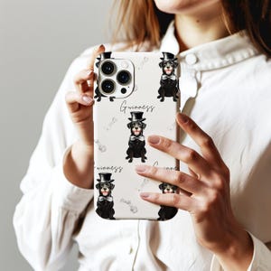 May include: A white phone case featuring a repeating pattern of black dogs wearing top hats and bow ties, with the word "Guinness" in script. The design also includes bone illustrations.