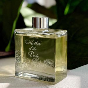 May include: A clear, square glass bottle with a silver cap, filled with a yellow liquid. The bottle is labeled "Luv and Lotus" and features the text "Mother of the Bride" with floral accents. The background is blurred green foliage.