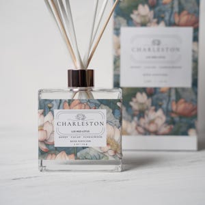 May include: A square glass reed diffuser with a metallic cap and white reeds. The label features a floral lotus pattern and text reading CHARLESTON LUV AND LOTUS HONEY CACAO SANDALWOOD REED DIFFUSER 4 oz | 113 g, matching the box behind it.