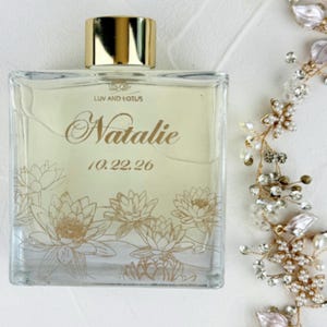 May include: A square glass perfume bottle with a gold cap, featuring the text LUV AND LOTUS, the name Natalie, and the date 10.22.26 in gold script. The bottle is decorated with gold line art of lotus flowers at the base.