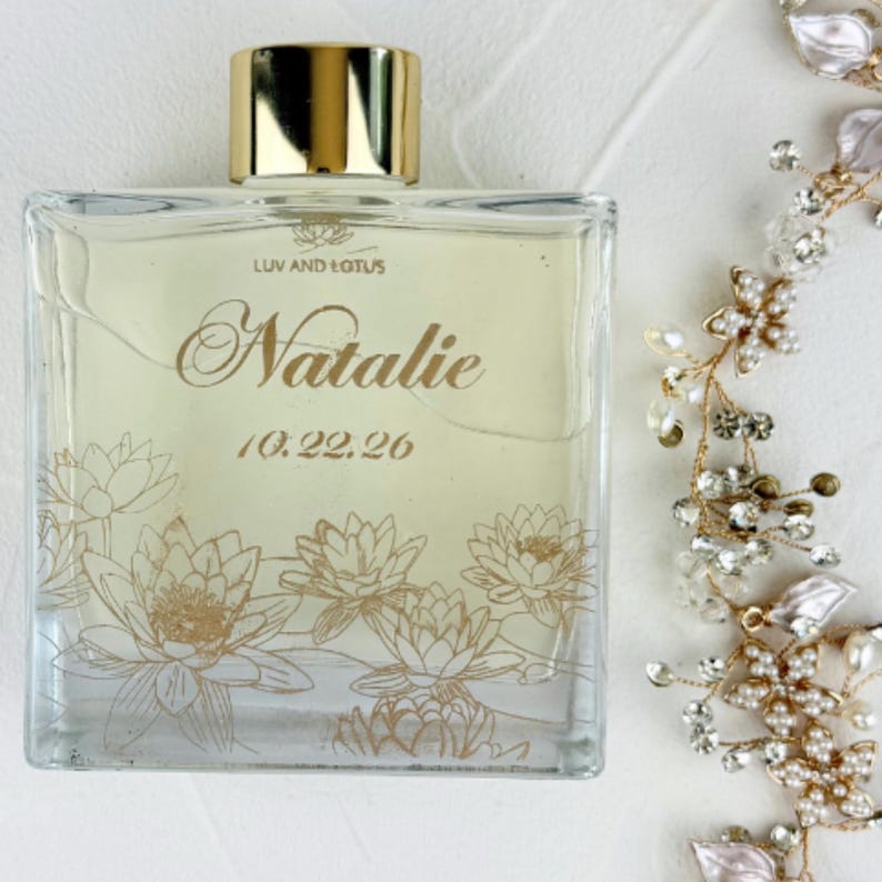May include: A square glass perfume bottle with a gold cap, featuring the text LUV AND LOTUS, the name Natalie, and the date 10.22.26 in gold script. The bottle is decorated with gold line art of lotus flowers at the base.