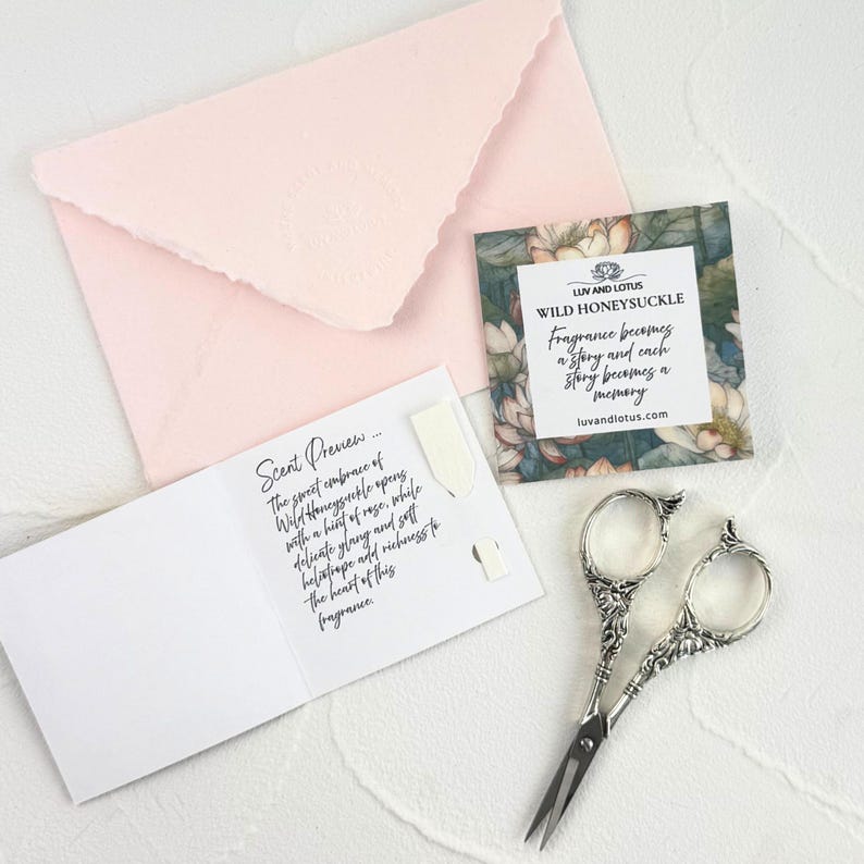 May include: A pale pink envelope with a deckled edge, a square card featuring a floral border and the text "LUV AND LOTUS WILD HONEYSUCKLE", a white folded card with "Scent Preview" text, and ornate silver-toned metal embroidery scissors.