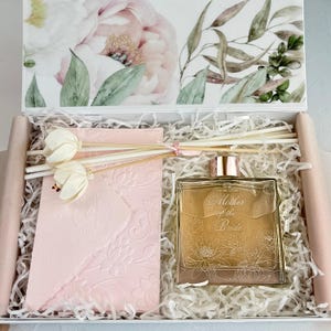 May include: A gift box containing a square glass bottle of gold-colored liquid with the text Mother of the Bride, a light pink embossed card, and two white floral reed diffuser sticks, all nestled in white paper shred with a floral lid.