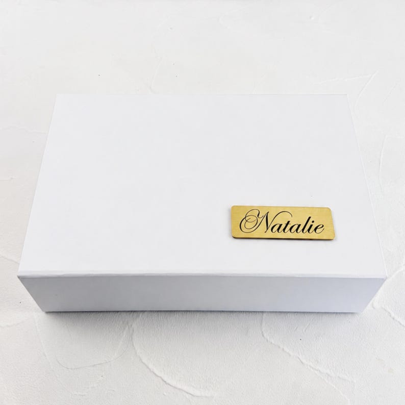 May include: A rectangular white gift box featuring a small, horizontal gold-toned metal plaque attached to the front. The plaque is engraved with the name Natalie in a black, elegant, cursive script font, centred on the gold surface.