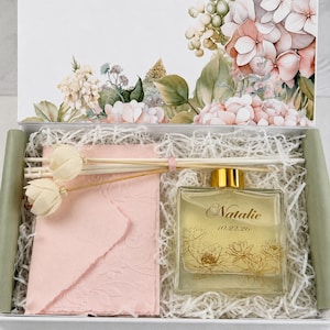 May include: A gift box containing a square glass diffuser bottle with a gold cap, labeled with the name Natalie and the date 10.22.26, featuring gold floral line art. Includes a pink textured envelope and white reed diffuser sticks.