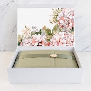 May include: A white rectangular gift box with an open lid featuring a watercolour illustration of pink and cream hydrangea flowers. Inside, the contents are wrapped in olive green paper secured with a matching ribbon and a gold circular seal.