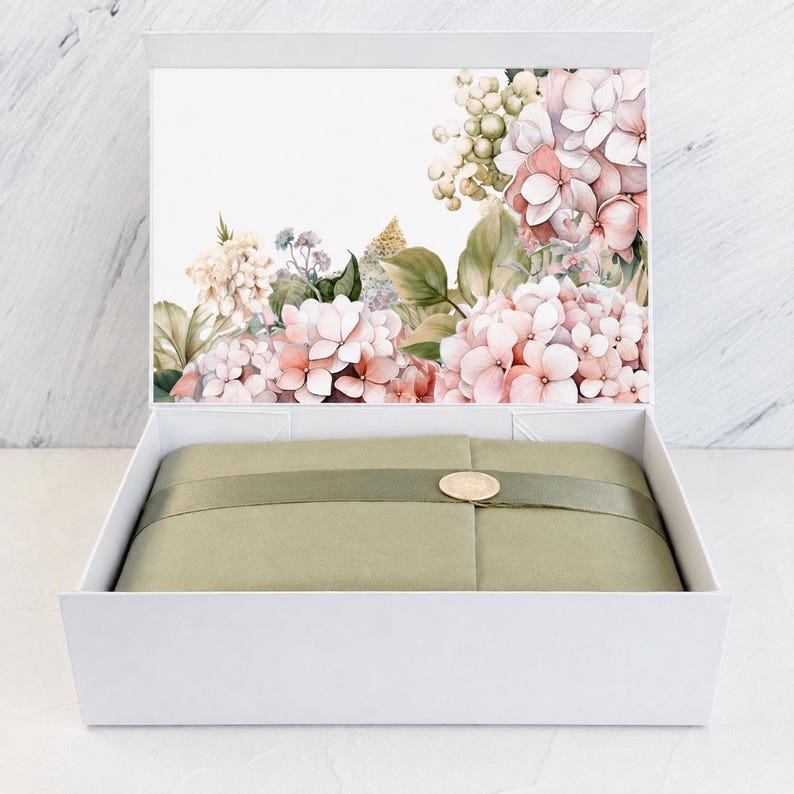 May include: A white rectangular gift box with an open lid featuring a watercolour illustration of pink and cream hydrangea flowers. Inside, the contents are wrapped in olive green paper secured with a matching ribbon and a gold circular seal.