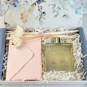 May include: A gift box containing a square glass perfume bottle with the text Maid of Honor 08.12.20, a light pink textured envelope, and two white floral reed diffuser sticks, all nestled in white paper shred filler with a floral lid.