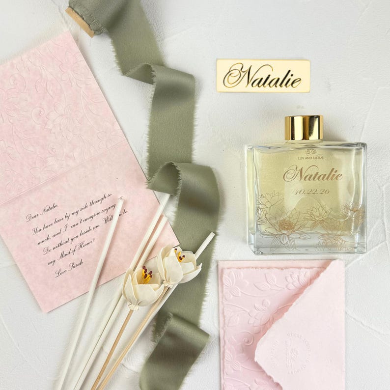 May include: A clear glass reed diffuser bottle with a gold cap, featuring the name Natalie and the date 10.22.26 printed on the front. Included are a pink textured card with handwritten text, a small gold nameplate, and sage green ribbon.