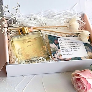 May include: A gift box with a perfume bottle, decorative sticks, and a card. The perfume bottle is gold and clear, with the name "Natalie" and the date "10.22.20" printed on it. The card reads "Will you be my Maid of Honor?"