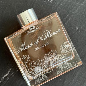 May include: A clear, square perfume bottle with a silver cap. The bottle is labeled "Luv and Lotus" and "Maid of Honor" with the date "08.12.26". White floral designs decorate the front of the bottle. The liquid inside has a light pink hue.