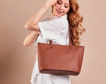 May include: A brown leather tote bag with a rectangular shape and two top handles. The bag is held by a person wearing a white shirt. The background is a solid beige color.