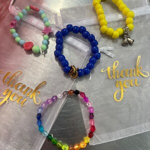 May include: Four colourful beaded bracelets in individual white organza bags with the words "thank you" printed in gold on the bags. The bracelets are made of blue, yellow, rainbow, and pastel beads.