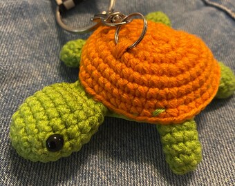 May include: A handmade crochet turtle keychain. The turtle has a bright orange shell and green head, feet, and tail. The keychain includes a silver metal ring and a black and white checkered strap.