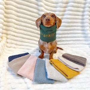 May include: A dachshund wearing a green sherpa fleece neck warmer with the name Franklin embroidered in gold thread, accompanied by a collection of similar sherpa fleece neck warmers in navy, beige, pink, blue, grey, mustard, white, and brown.