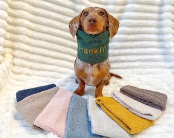 May include: A dachshund wearing a green sherpa fleece neck warmer with the name Franklin embroidered in gold thread, accompanied by a collection of similar sherpa fleece neck warmers in navy, beige, pink, blue, gray, mustard, white, and brown.