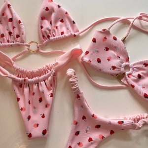 May include: A pink bikini with a strawberry print. The top is a triangle style with adjustable straps and a ring detail. The bottom is a cheeky style with a high leg cut.