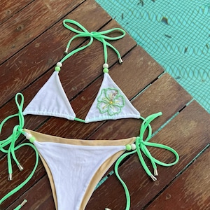 Puede incluir: Bikini blanco con tirantes verdes y un adorno floral en la parte superior. El bikini está sobre una superficie de madera.