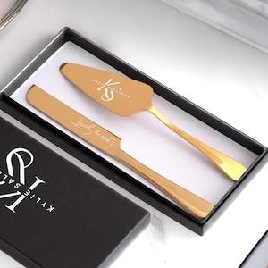 May include: A black box with a gold cake server and knife set. The box has the text "KYLIE & SALIMAN" and the initials "KS" on it. The server and knife are engraved with the text "Love Sweet" and "KS".