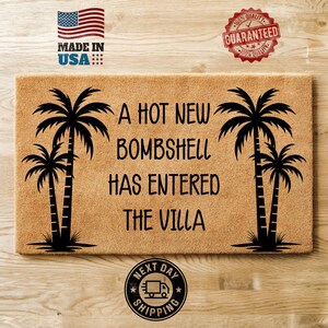May include: A rectangular tan coir doormat featuring black text that reads A HOT NEW BOMBSHELL HAS ENTERED THE VILLA. The design includes black silhouettes of two palm trees on the left side and two palm trees on the right side of the text.