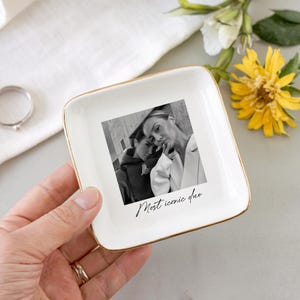 Peut inclure: Un vide-poche carr&eacute; en c&eacute;ramique blanche avec un bord dor&eacute;, orn&eacute; d'une photo en noir et blanc et du texte "Most iconic duo". Le vide-poche est tenu dans une main, avec une bague visible en arri&egrave;re-plan.