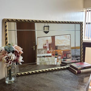 May include: A rectangular mirror with a gold, ornate frame. The mirror is divided into sections and reflects a room with a painting and a vase of flowers. The frame has a detailed, textured design.