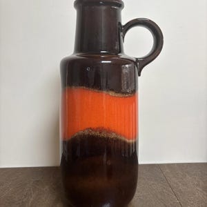 May include: A tall ceramic vase with a dark brown top and base, and a vibrant orange band in the middle. The vase has a handle and a textured surface. This decorative piece is suitable for home decor.