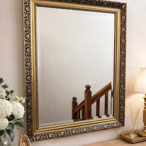 May include: Rectangular mirror with a gold ornate frame. The frame has a dark brown decorative pattern. The mirror is hanging on a light beige wall. A wooden table with a lamp and flowers is in front of the mirror.