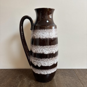 May include: A tall, brown ceramic vase with a handle. The vase has horizontal bands of white and brown, creating a textured, striped pattern. The vase has a glossy finish and a wide opening, suitable for displaying flowers or as a decorative piece.