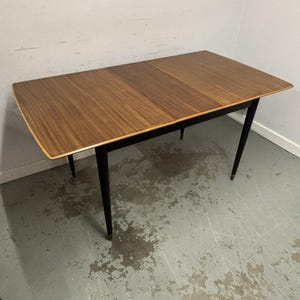 May include: A mid-century dining table with a rectangular top and four black legs. The tabletop is a warm brown with a wood grain pattern. The legs are slender and angled, with brass-coloured tips.