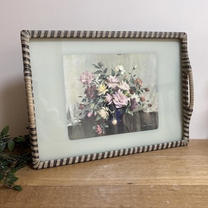 May include: A framed floral print with a rectangular shape. The frame has a woven, striped design in brown and beige. The print depicts a bouquet of pink, white, and yellow flowers in a blue vase. The background is a soft, muted colour.