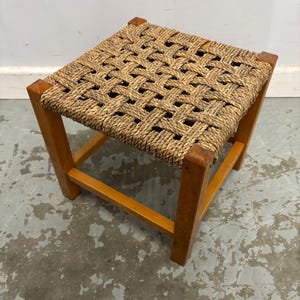 May include: A small, square wooden stool with a woven rope seat. The frame is made of light brown wood, and the seat is a natural tan colour. The stool is sturdy and appears to be handmade. It is a simple, functional piece of furniture.