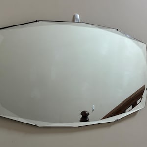 May include: An oval mirror with a black frame and beveled edges. The mirror is mounted on a light-coloured wall. The reflection shows a wooden railing and a dark-coloured object.