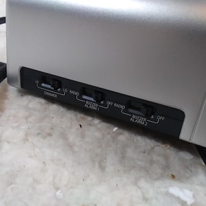 May include: Close-up of a silver clock radio with black buttons and labels. The buttons are labelled "Dimmer", "Lo", "Radio", "Buzzer Alarm 1", "Off Radio", "Buzzer Alarm 2", and "Off".