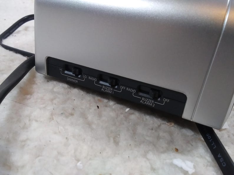 May include: Close-up of a silver clock radio with black buttons and labels. The buttons are labelled "Dimmer", "Lo", "Radio", "Buzzer Alarm 1", "Off Radio", "Buzzer Alarm 2", and "Off".