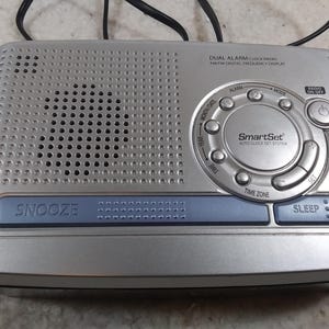 May include: Silver dual alarm clock radio with a digital display and a SmartSet auto clock set system. The radio has buttons for snooze, sleep, time zone, set, mode, and alarm.